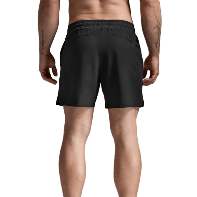 Rear view of a man wearing black athletic shorts, white socks, and white sneakers against a gray studio background.