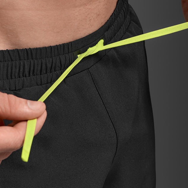 Close-up of a person wearing black athletic shorts and adjusting a bright neon yellow drawstring at the elastic waistband.