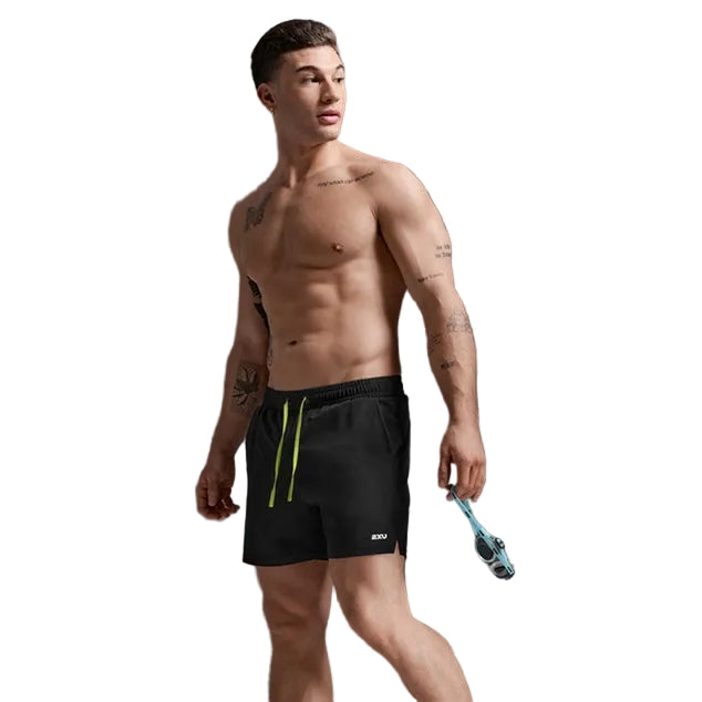 A shirtless male model wearing black athletic shorts with bright green drawstrings, standing barefoot against a dark gray studio background and holding a pair of blue swim goggles.