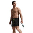 A shirtless male model wearing black athletic shorts with bright green drawstrings, standing barefoot against a dark gray studio background and holding a pair of blue swim goggles.