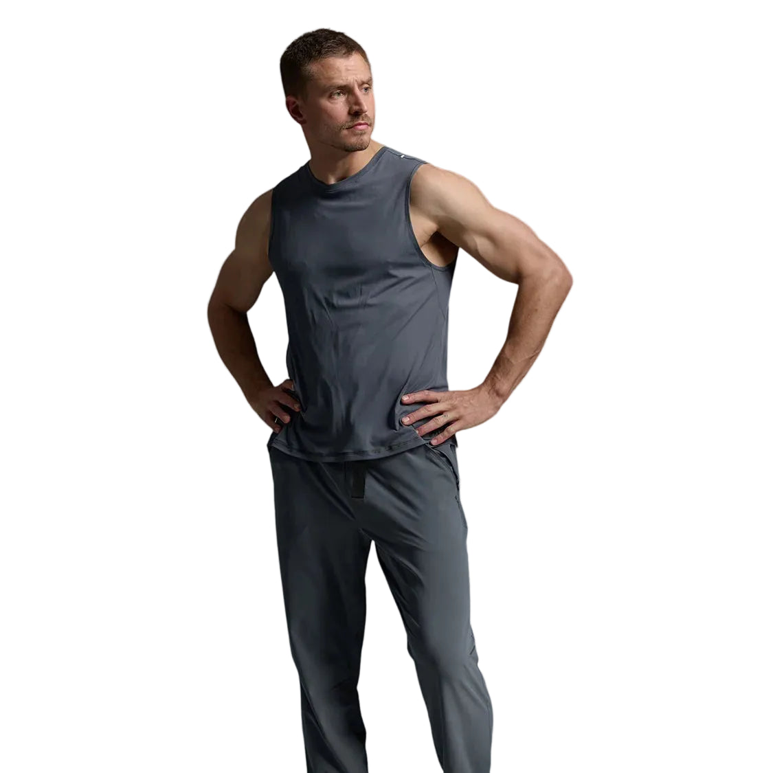 A man modeling a dark gray sleeveless athletic top and matching gray jogger pants, paired with beige sneakers, against a light gray studio background.