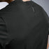 Close-up back view of a person wearing a black textured athletic short-sleeve shirt with a small vertical logo near the upper neckline, against a light gray studio background.