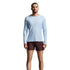 A man modeling a light blue long-sleeve athletic shirt and dark brown shorts against a light gray studio background.