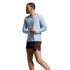 A man in a light blue long-sleeve athletic shirt, dark brown shorts, white socks, and white running shoes is shown in profile against a light gray studio background.