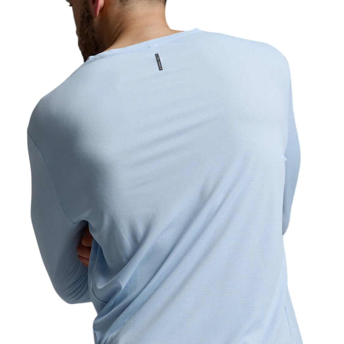 A man shown from the back wearing a light blue long-sleeve athletic shirt and dark brown pants against a gray studio background.