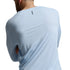 A man shown from the back wearing a light blue long-sleeve athletic shirt and dark brown pants against a gray studio background.