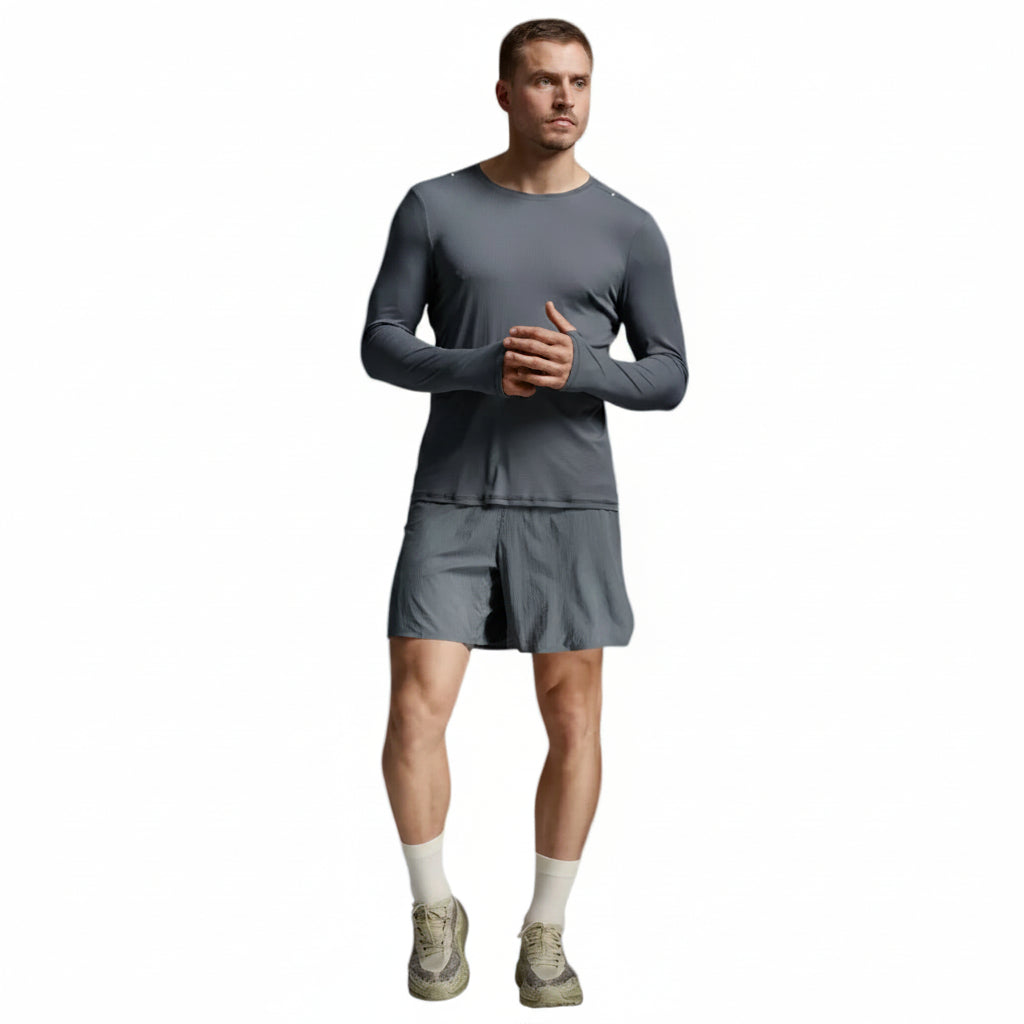 A man modeling a dark gray long-sleeve athletic shirt and matching shorts, paired with off-white socks and running shoes, against a light gray studio background.