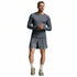 A man modeling a dark gray long-sleeve athletic shirt and matching shorts, paired with off-white socks and running shoes, against a light gray studio background.