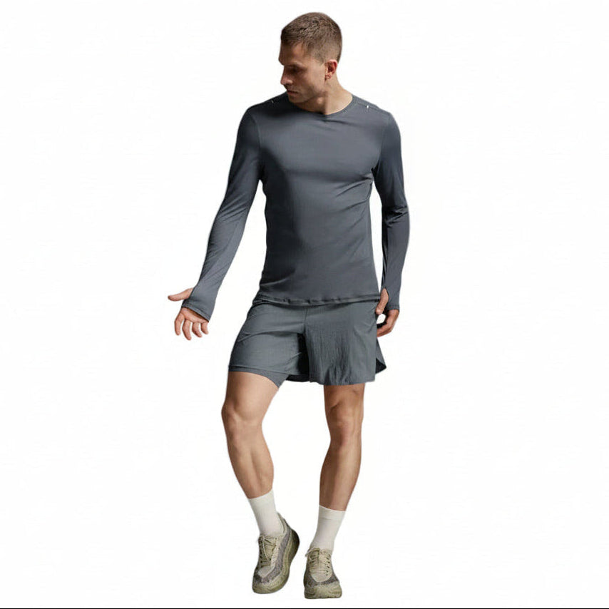 A man modeling a dark gray long-sleeve athletic shirt and matching gray shorts, paired with off-white socks and gray running shoes, against a light gray studio background.