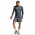 A man modeling a dark gray long-sleeve athletic shirt and matching gray shorts, paired with off-white socks and gray running shoes, against a light gray studio background.