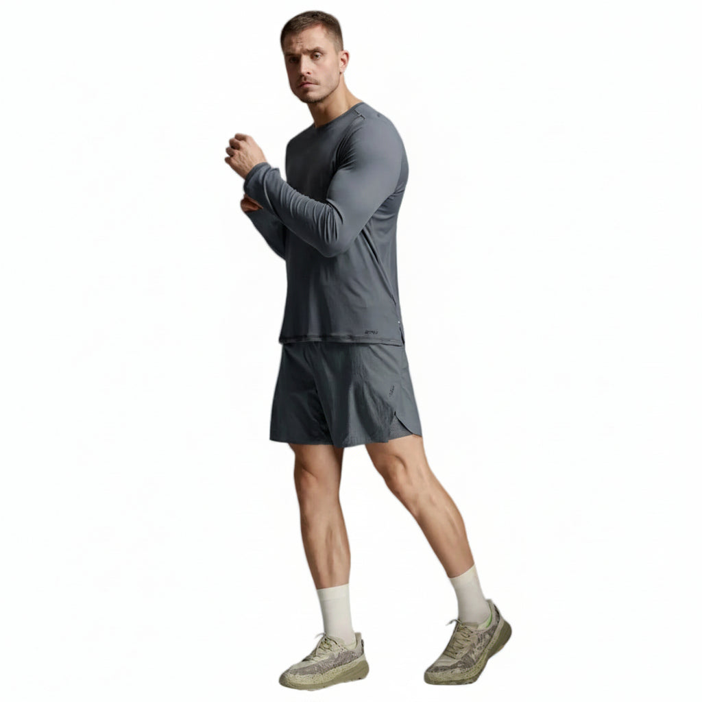 A man modeling a dark gray long-sleeve athletic shirt and matching shorts, paired with off-white socks and beige sneakers, against a light gray studio background.