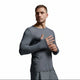 A man wearing a tight-fitting long-sleeve athletic shirt in dark gray, posed against a neutral light gray studio background.