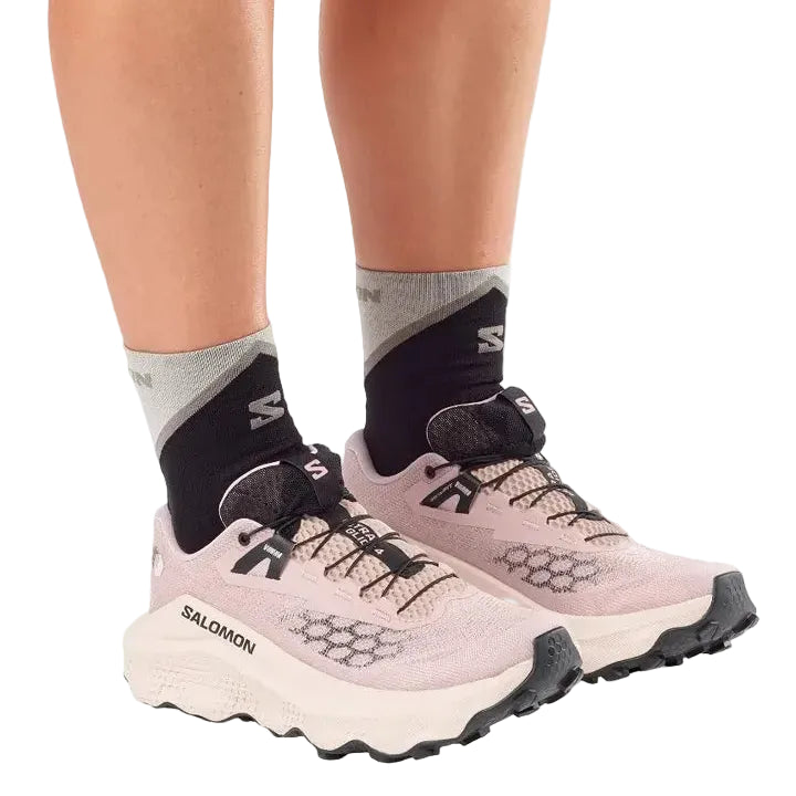 Salomon trail running shoes in a light pink and white colorway, shown on a model wearing black and gray Salomon socks against a white background.