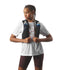 A woman wearing a white t-shirt and dark brown bike shorts models a black Salomon running hydration vest with two water bottles, set against a plain light gray background.