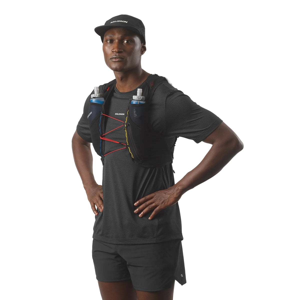 A man wearing a black athletic t-shirt, black shorts, and a black cap, modeling a black hydration vest with red and yellow straps and two water bottles, against a light gray background.