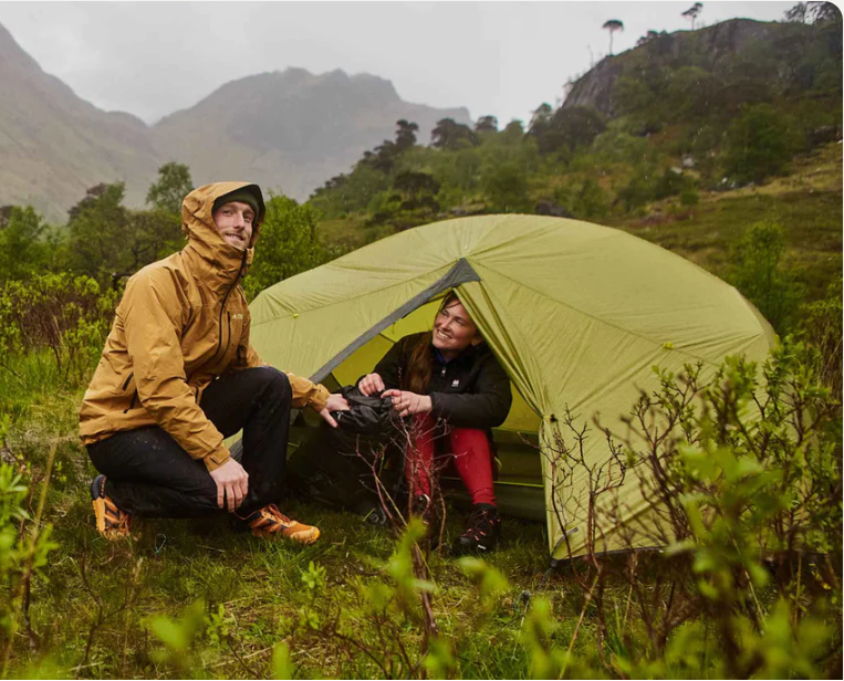 Two people camping in a green tent in a mountainous, forested area.