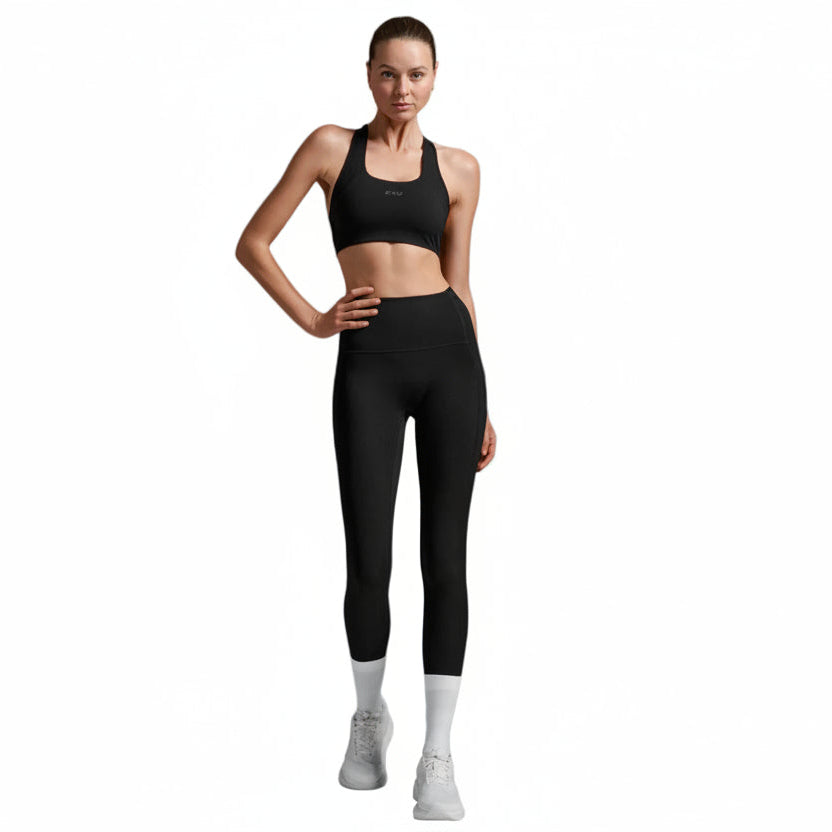 A woman modeling a black sports bra and matching black high-waisted leggings with white socks and white sneakers, standing against a solid white background.