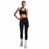 A woman modeling a black sports bra and matching black high-waisted leggings with white socks and white sneakers, standing against a solid white background.