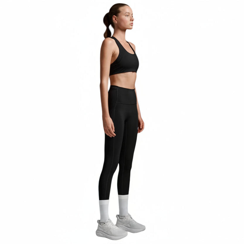 A woman in a black sports bra and black high-waisted leggings stands in profile against a light gray studio background, wearing white socks and light gray sneakers.