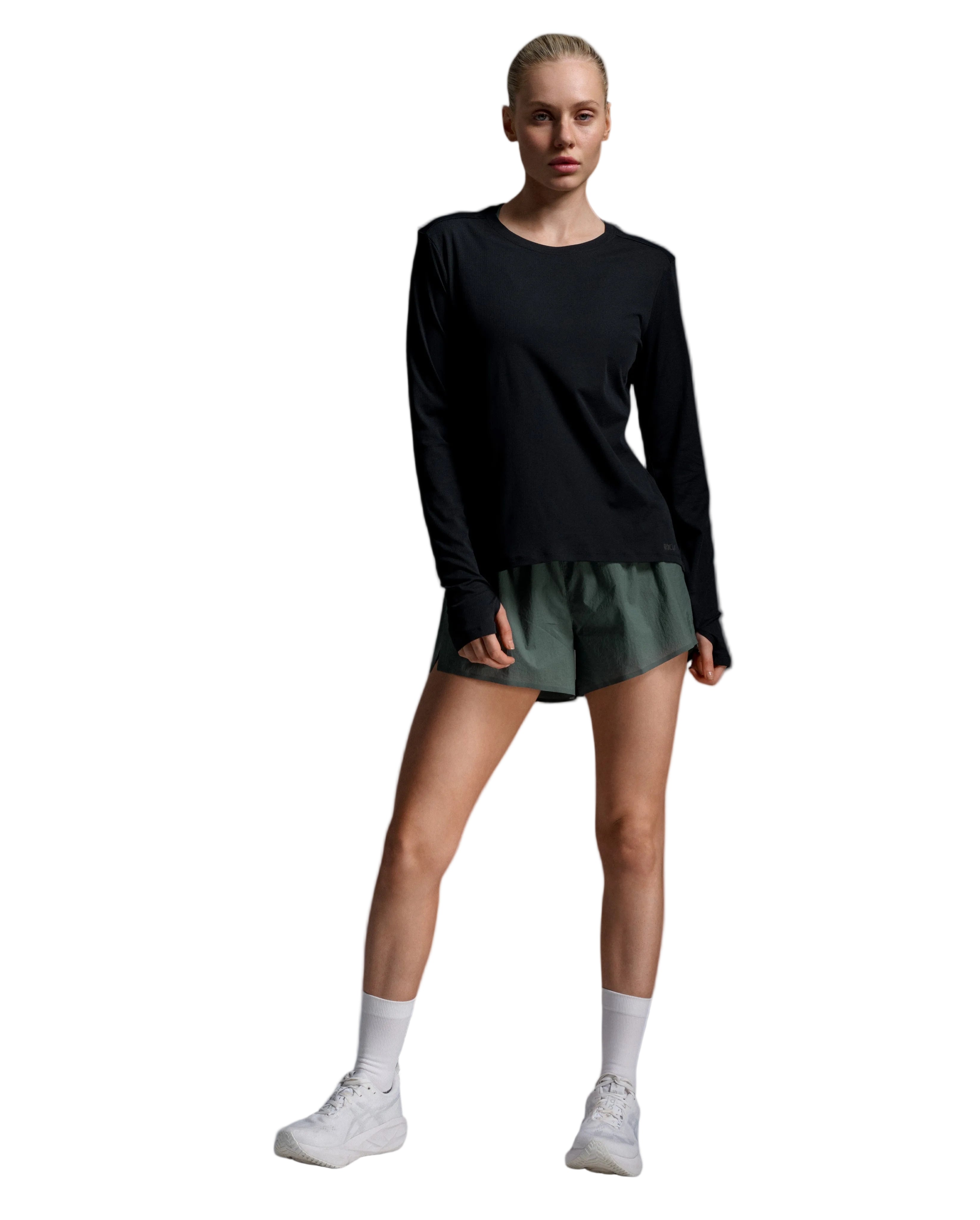 A woman modeling a black long-sleeve athletic top and dark green athletic shorts, paired with white socks and white sneakers, against a light gray studio background.