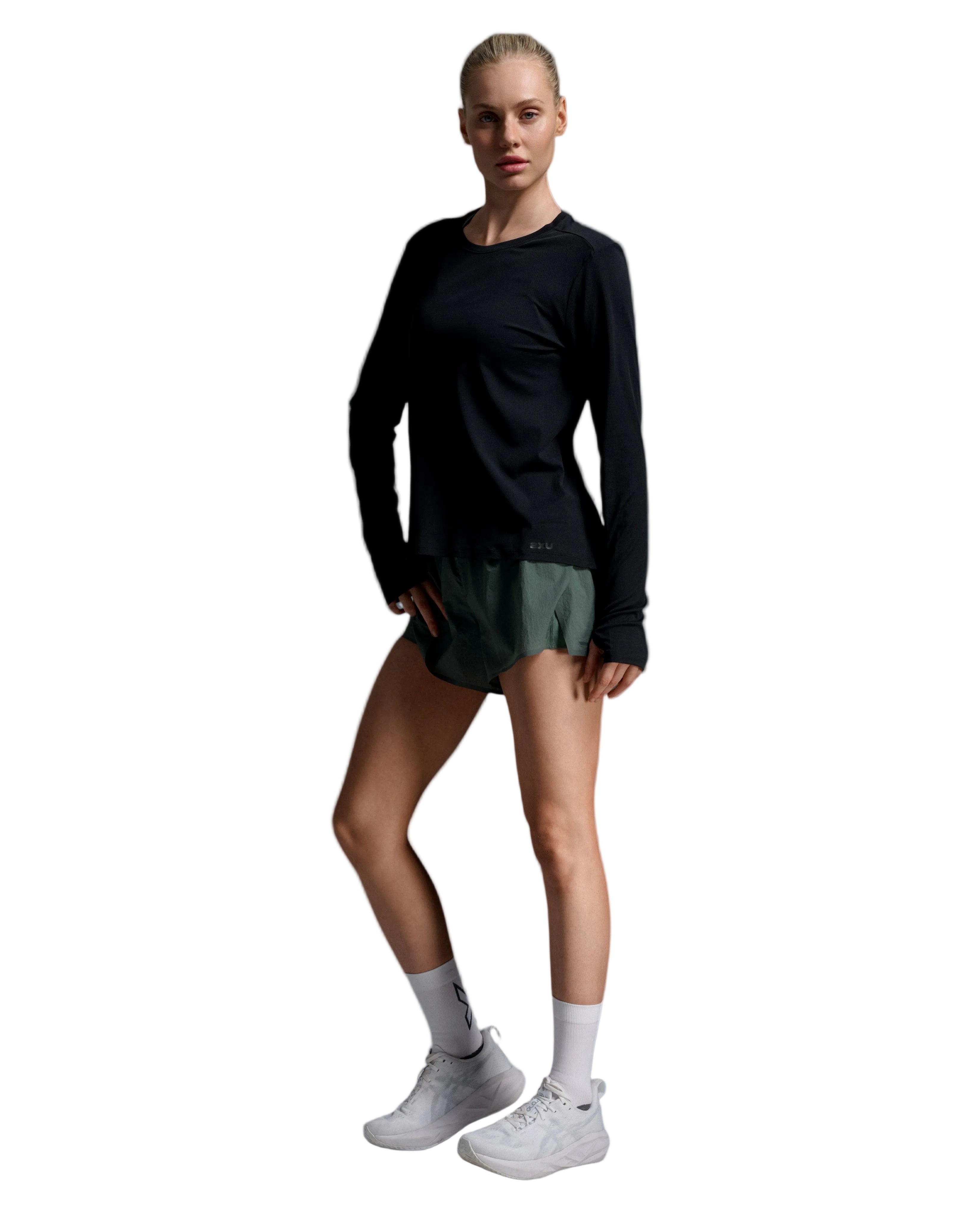 A female model stands in a studio wearing a black long-sleeve athletic top, dark green athletic shorts, white crew socks, and white sneakers against a light gray gradient background.