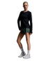 A female model stands in a studio wearing a black long-sleeve athletic top, dark green athletic shorts, white crew socks, and white sneakers against a light gray gradient background.