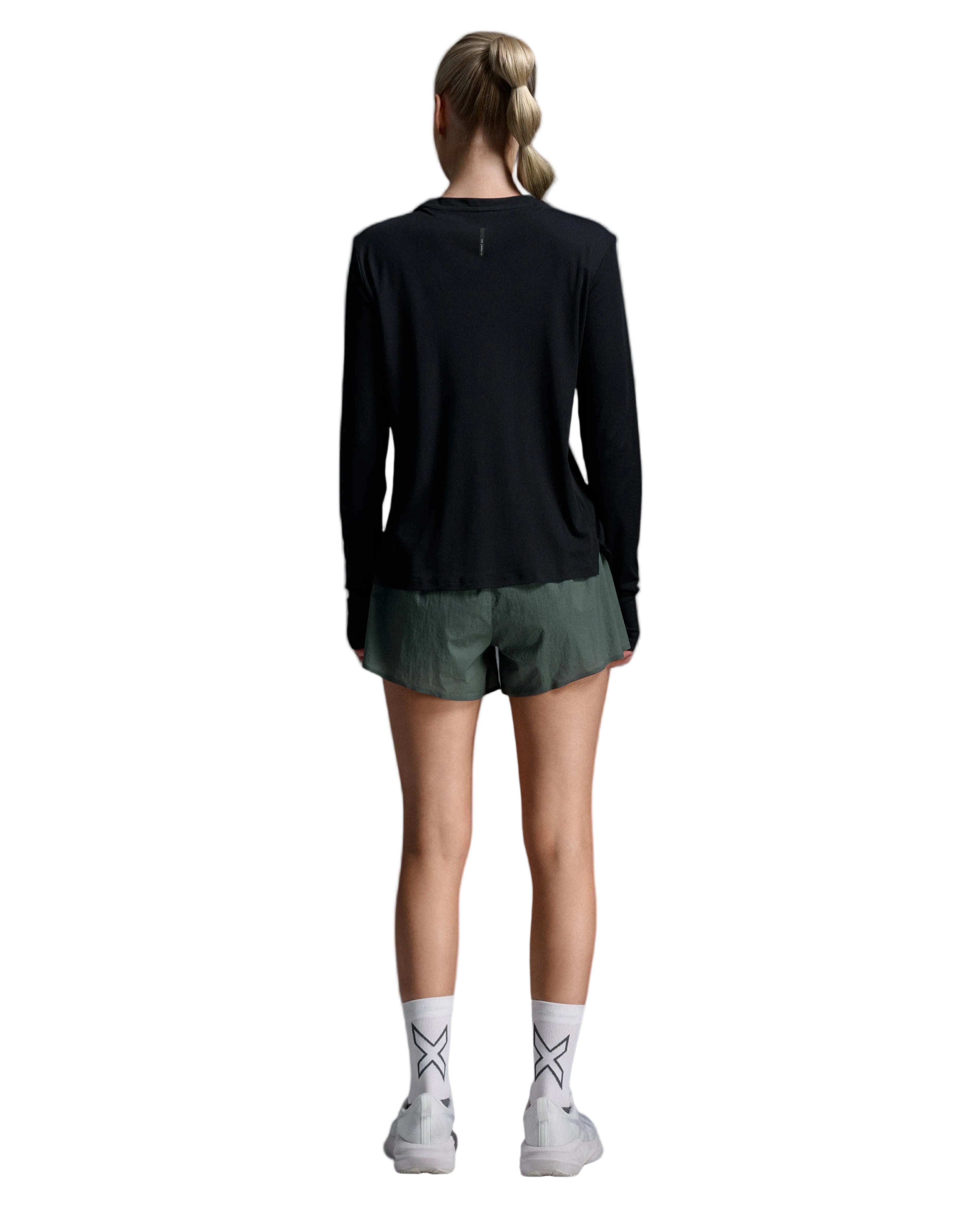 A woman is shown from behind wearing a black long-sleeve athletic top, olive green running shorts, white crew socks with a black geometric pattern, and white sneakers, set against a light gray studio background.