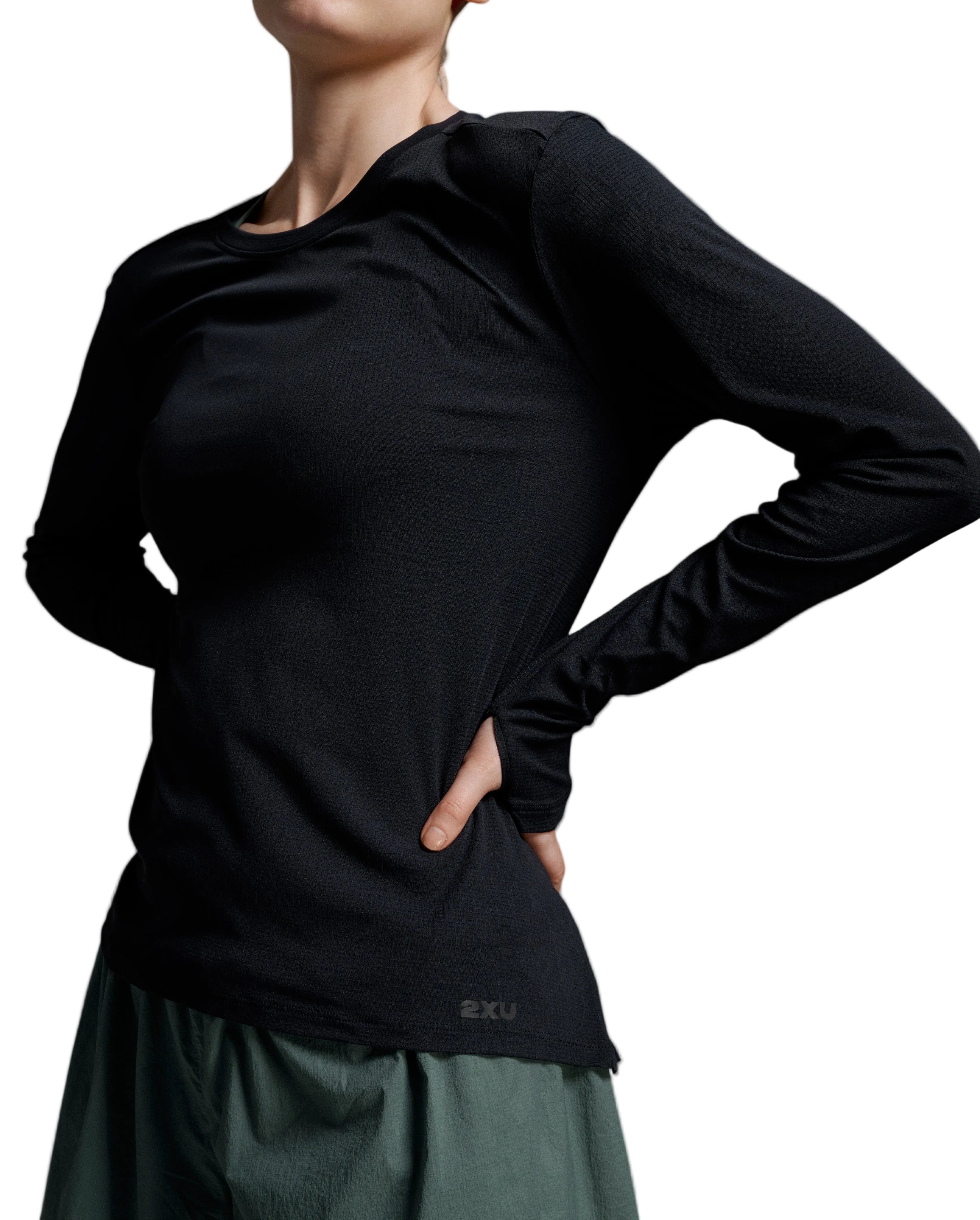 A close-up of a model wearing a black long-sleeve athletic top with a small '2XU' logo near the hem, paired with a dark green skirt, against a neutral gray studio background.
