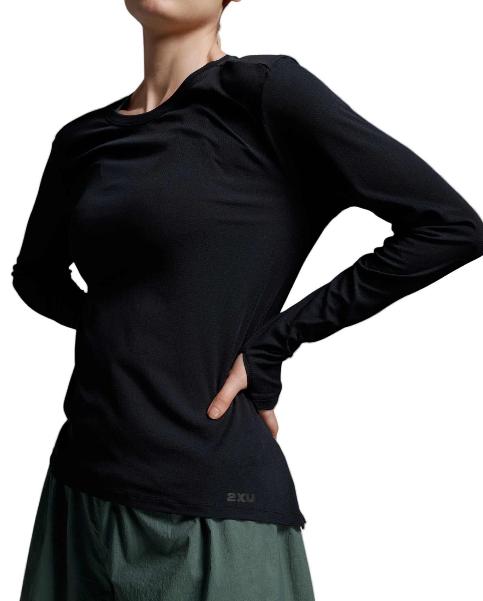 A close-up of a model wearing a black long-sleeve athletic top with a small '2XU' logo near the hem, paired with a dark green skirt, against a neutral gray studio background.