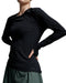 A close-up of a model wearing a black long-sleeve athletic top with a small '2XU' logo near the hem, paired with a dark green skirt, against a neutral gray studio background.