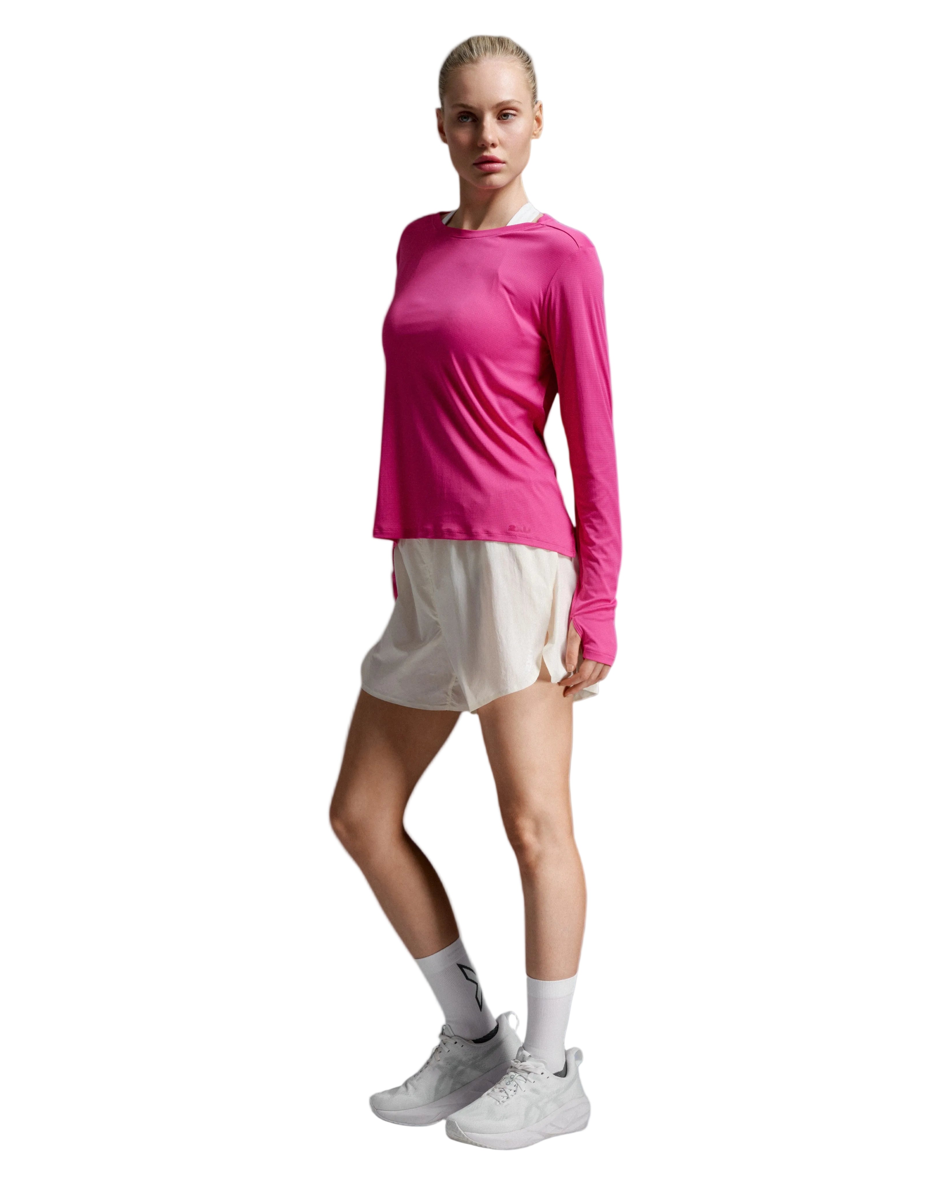 A woman modeling a bright pink long-sleeve athletic top paired with light beige shorts, white socks, and white sneakers against a light gray studio background.