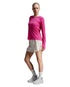A woman modeling a bright pink long-sleeve athletic top paired with light beige shorts, white socks, and white sneakers against a light gray studio background.