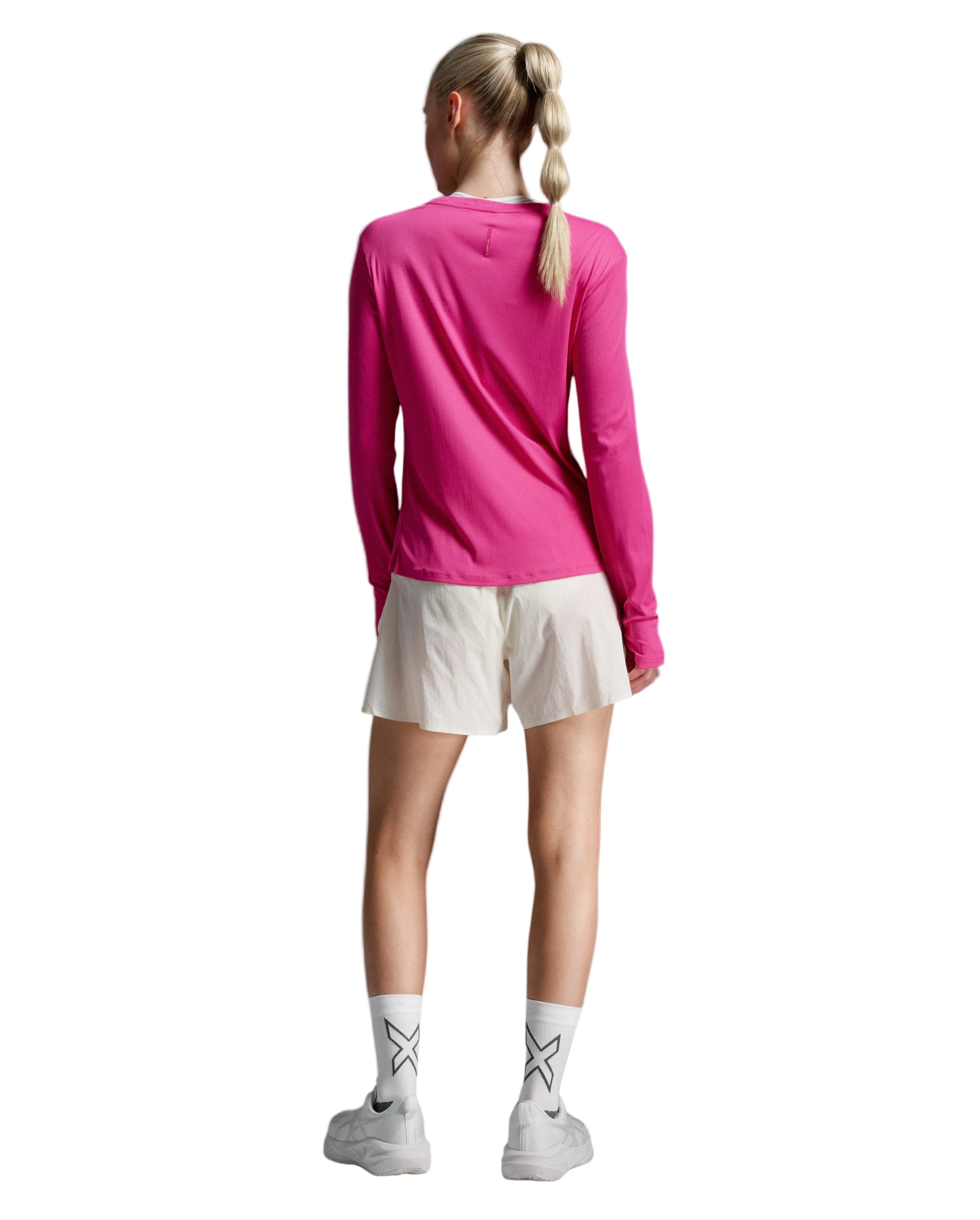 A woman shown from the back wearing a bright pink long-sleeve athletic top, off-white athletic shorts, white socks, and white sneakers against a light gray studio background.