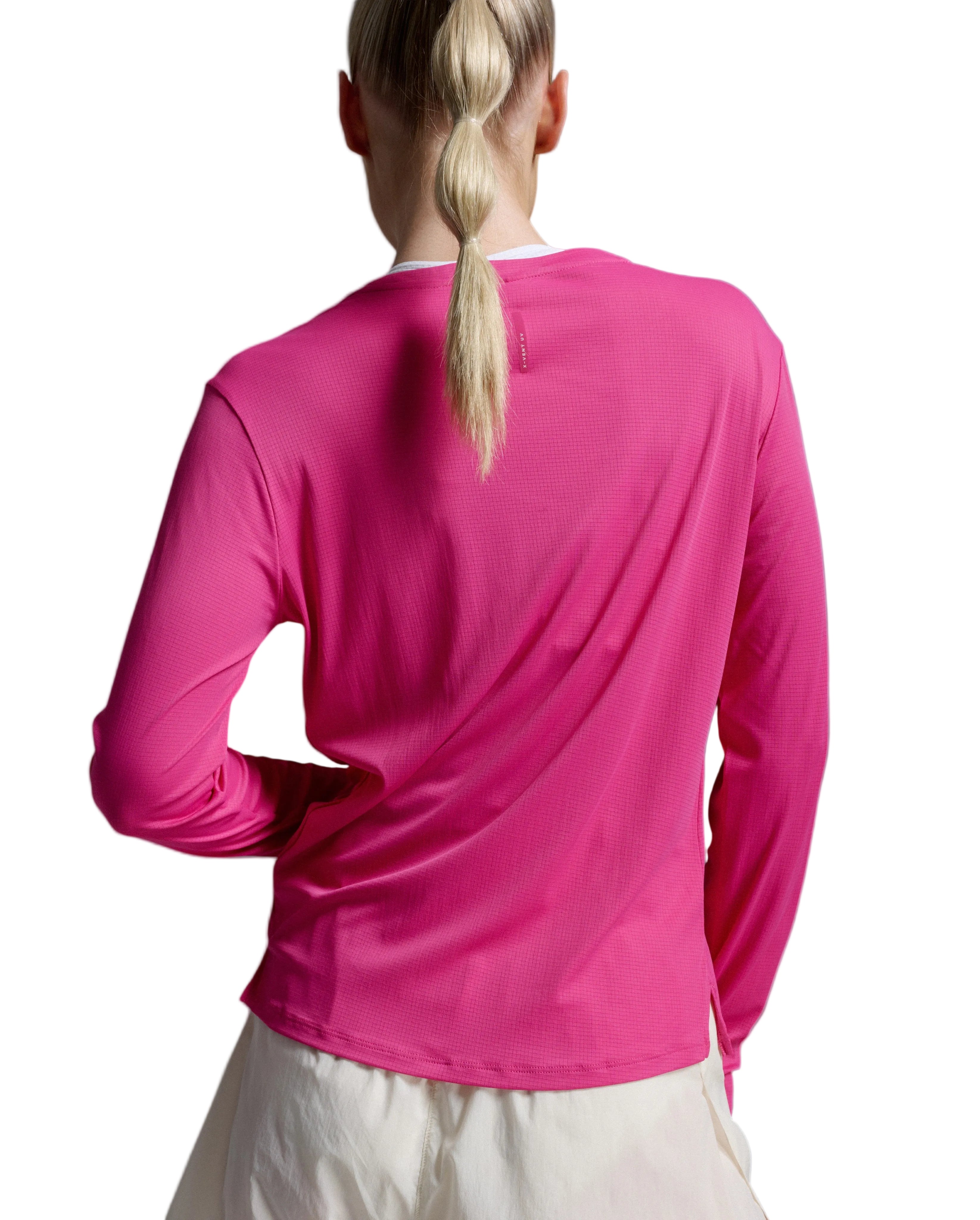 A woman viewed from behind wearing a bright pink long-sleeve athletic top and light-colored bottoms against a light gray studio background.