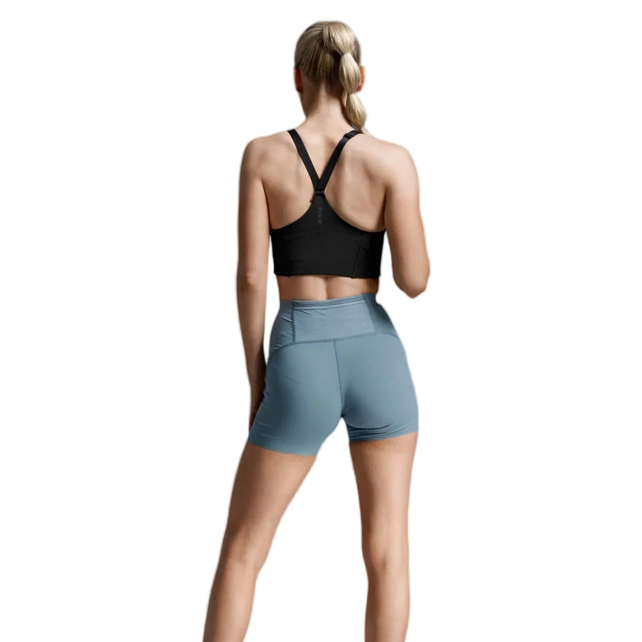 A woman shown from behind wearing a black sports bra and light blue athletic shorts, standing against a light gray studio background.